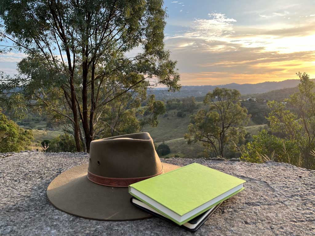 An Akubra hat on a rock, with two notebooks leaning on the brim. There is a gorgeous view over rural valleys, with a large gum tree to the left of the picture. An orange sunset is to the right of the picture