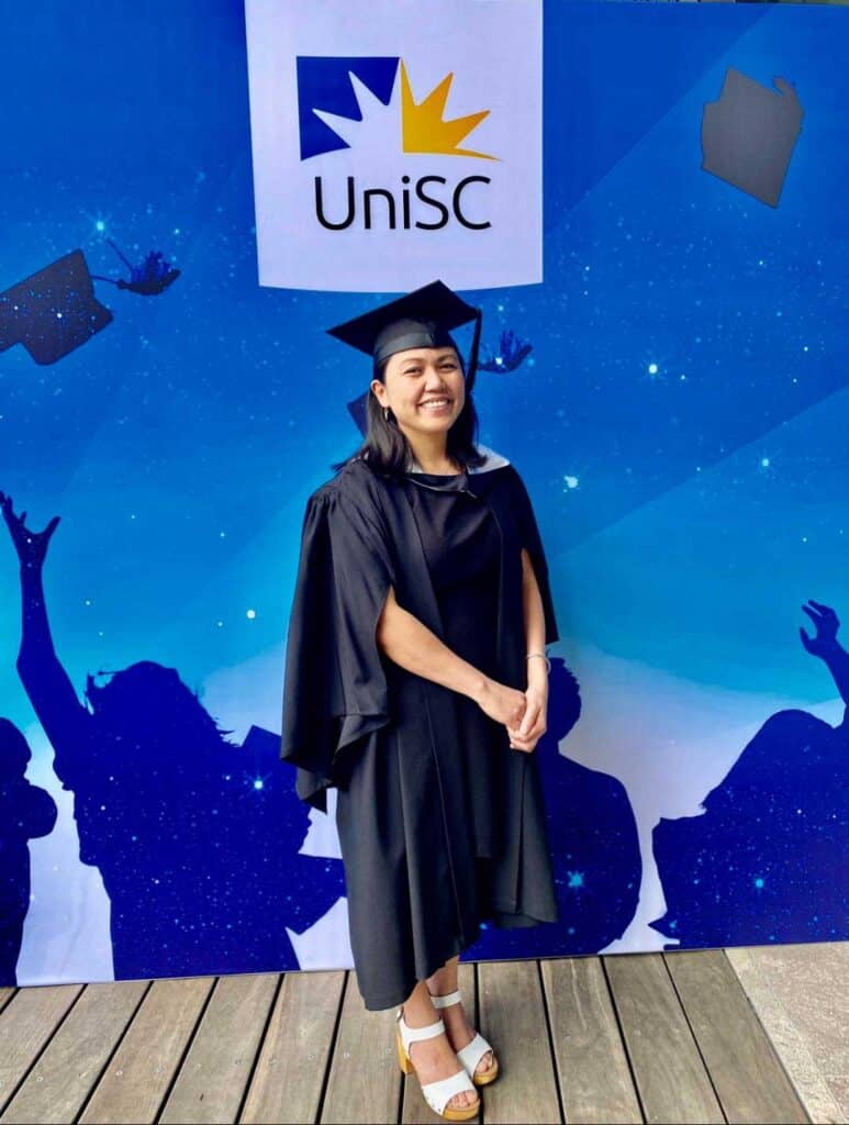 Mheemhar Kennedy in her graduating cap and gown against a blue wall with UniSC behind her.